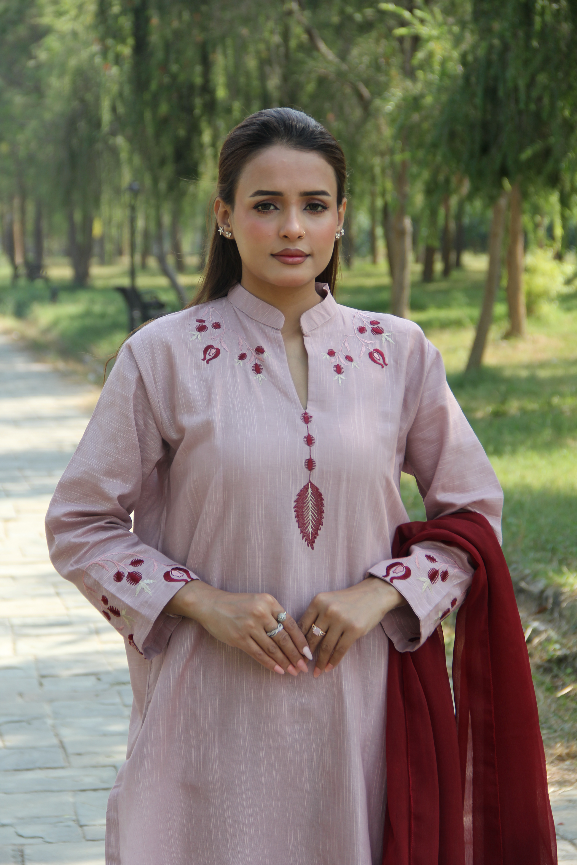 Woman in a light pink traditional outfit with red embroidery standing outdoors.