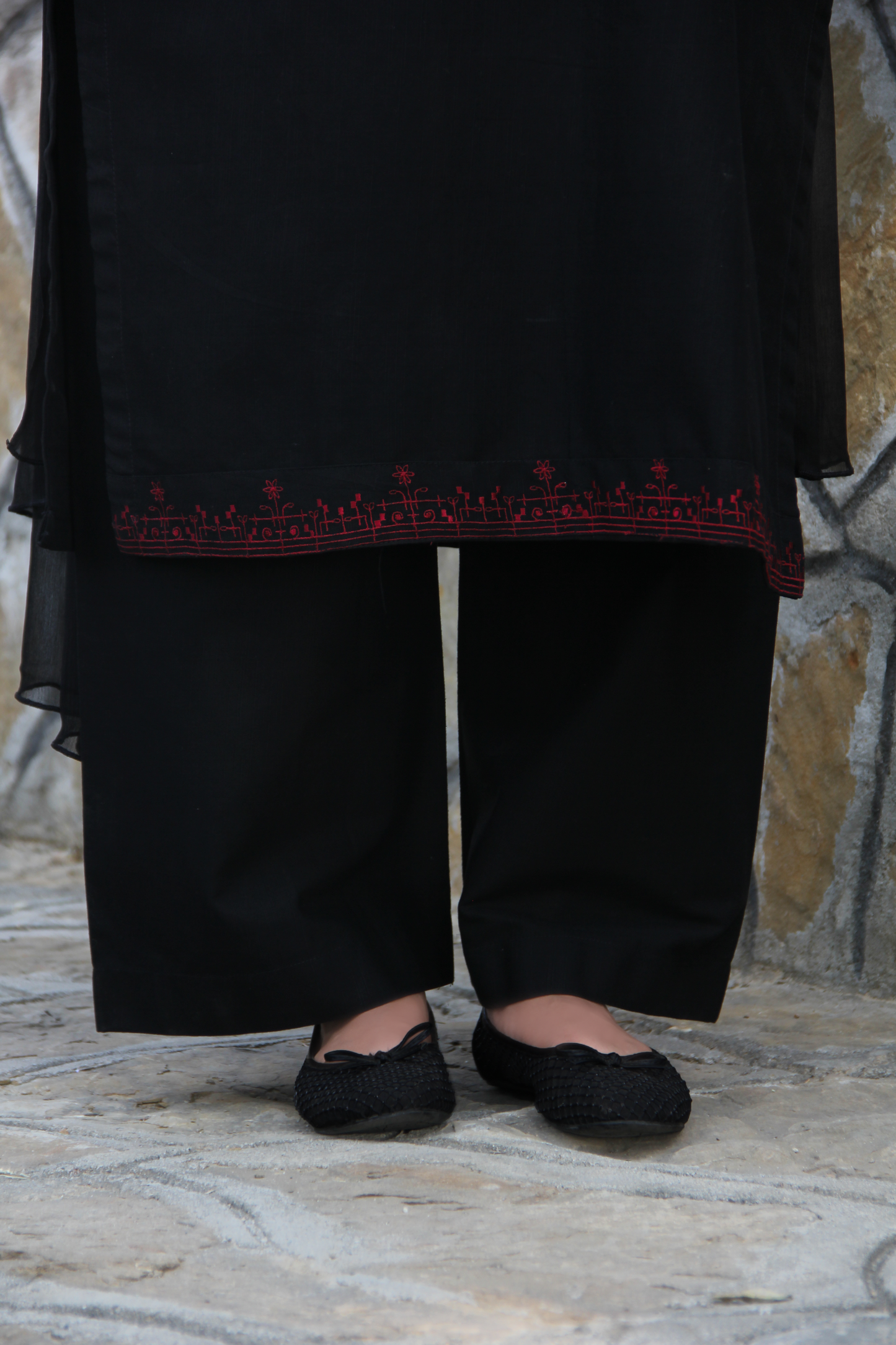 Person wearing a black outfit with red embroidery on a stone floor.