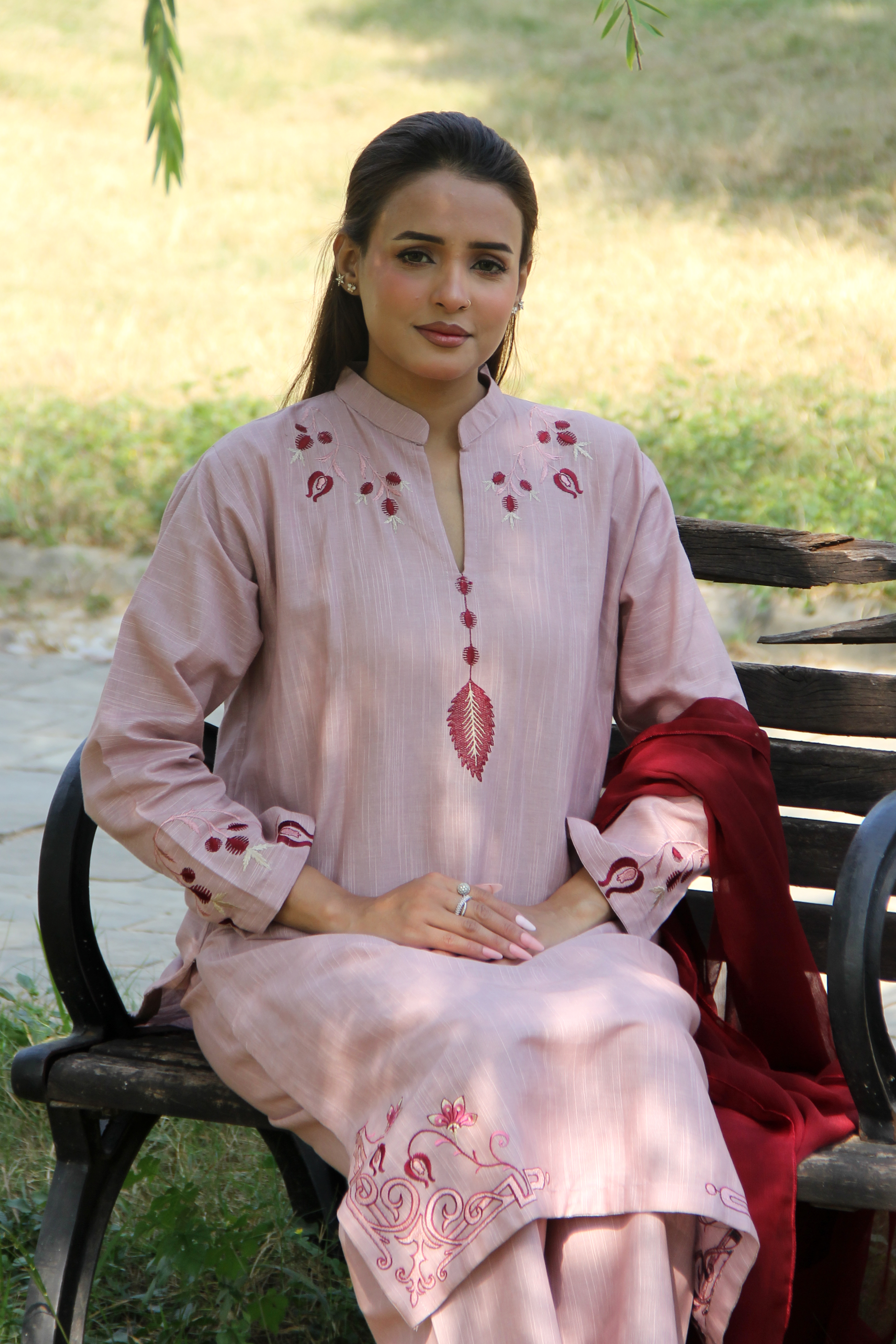 Woman in a light pink embroidered traditional outfit sitting on a bench outdoors.