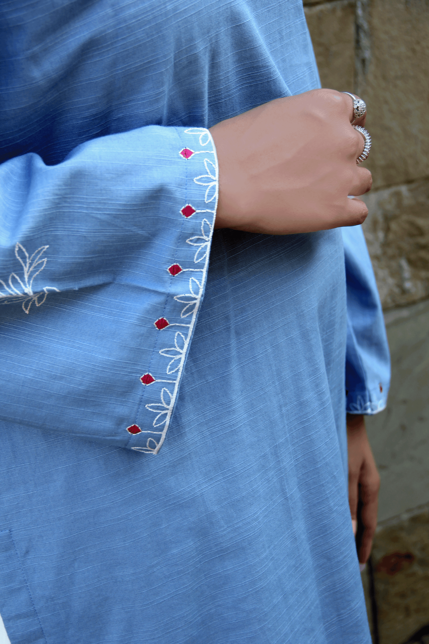 Close-up of a blue garment with intricate white and red embroidery on a stone wall background