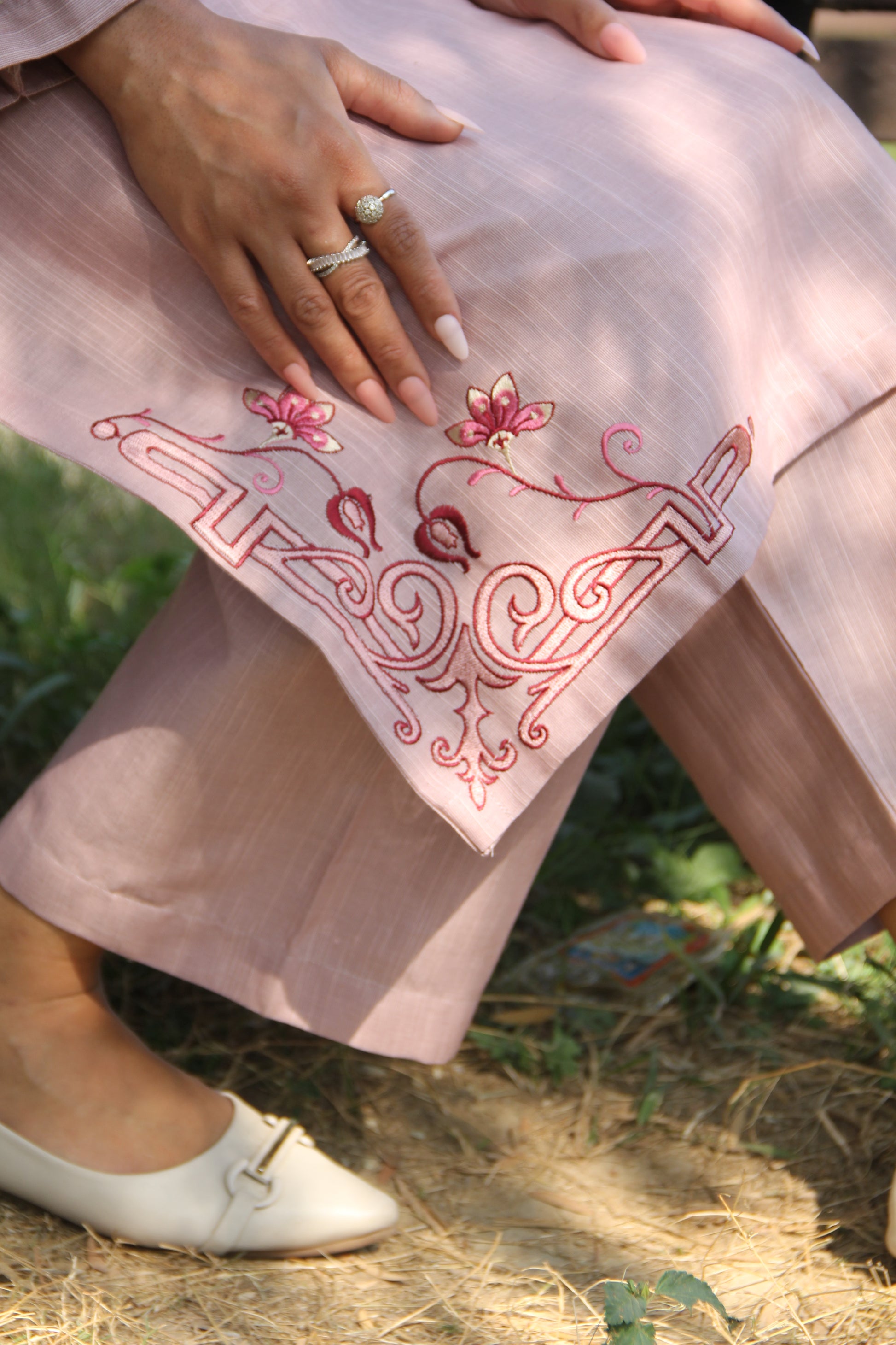 Person wearing a light pink dress with intricate red embroidery, sitting on a wooden bench outdoors.