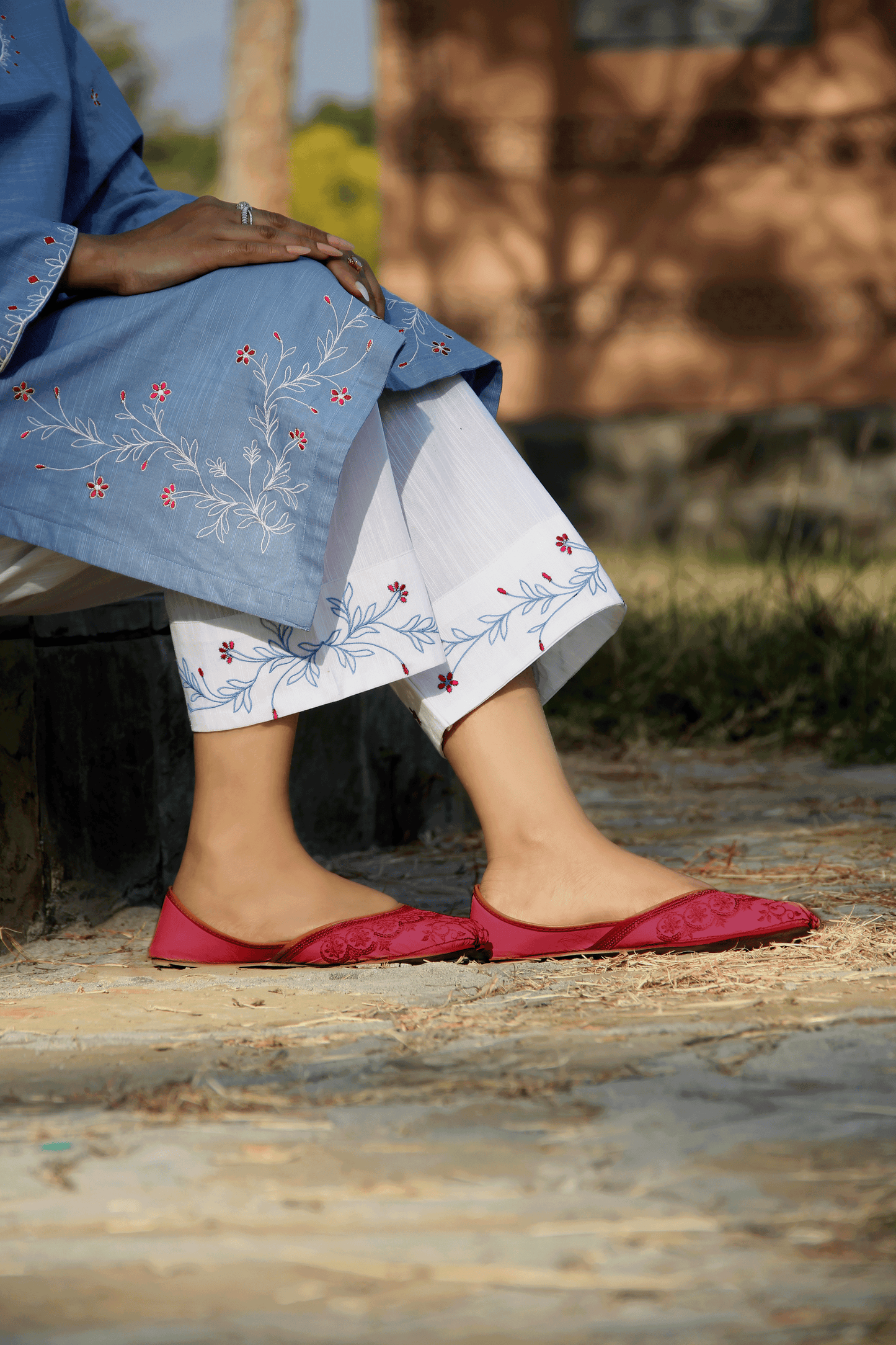 Person wearing a blue embroidered outfit with white pants and red shoes, sitting outdoors.