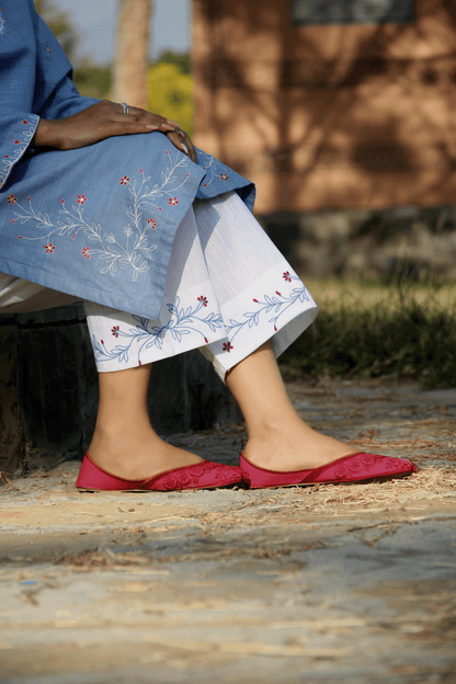 Person wearing a blue embroidered outfit with white pants and red shoes, sitting outdoors.