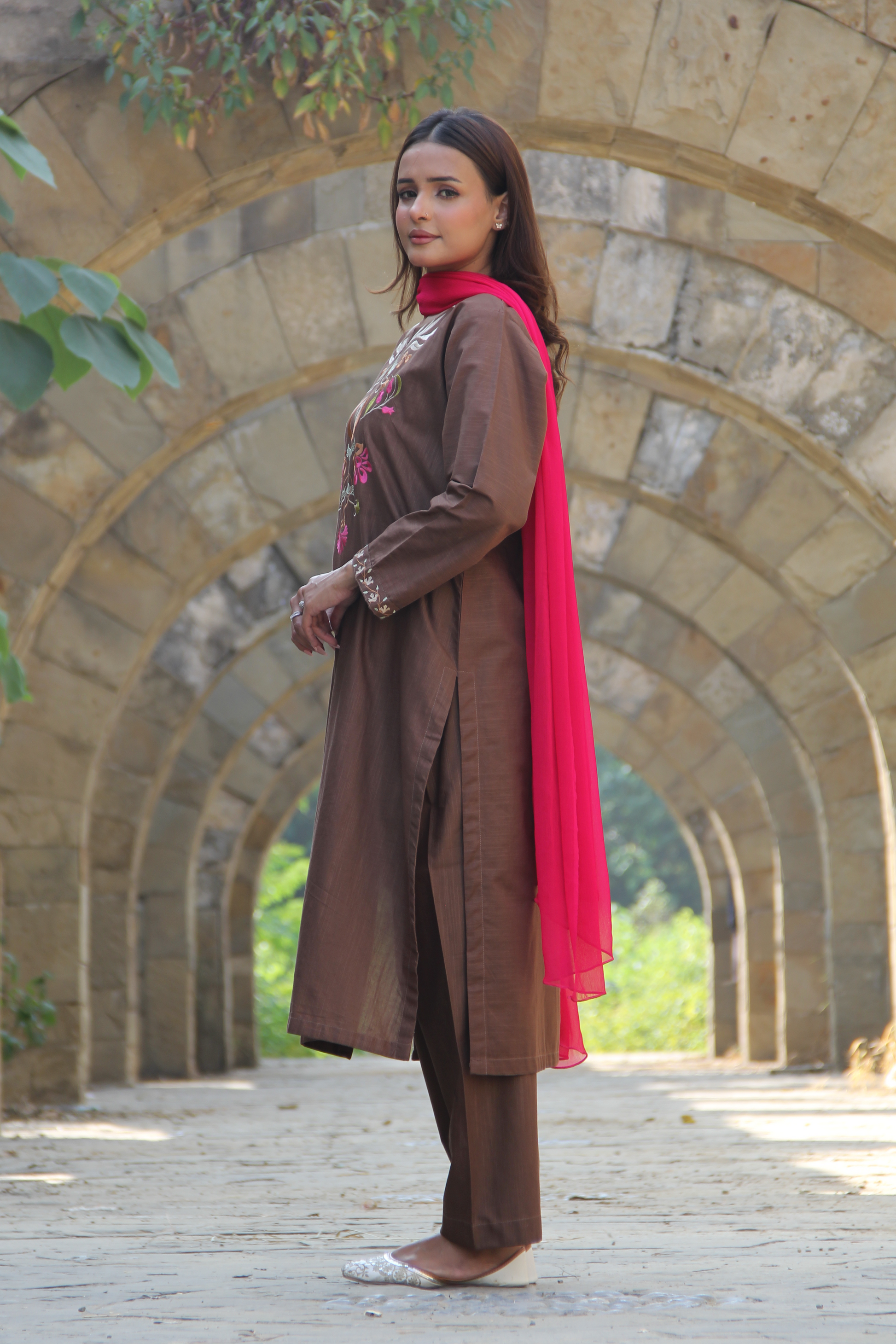 Woman in brown traditional outfit with red scarf standing in front of stone arches.