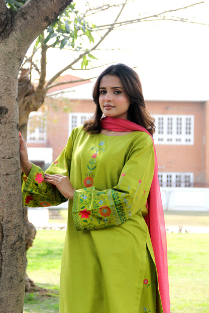 Woman in a green traditional outfit with floral patterns standing next to a tree.