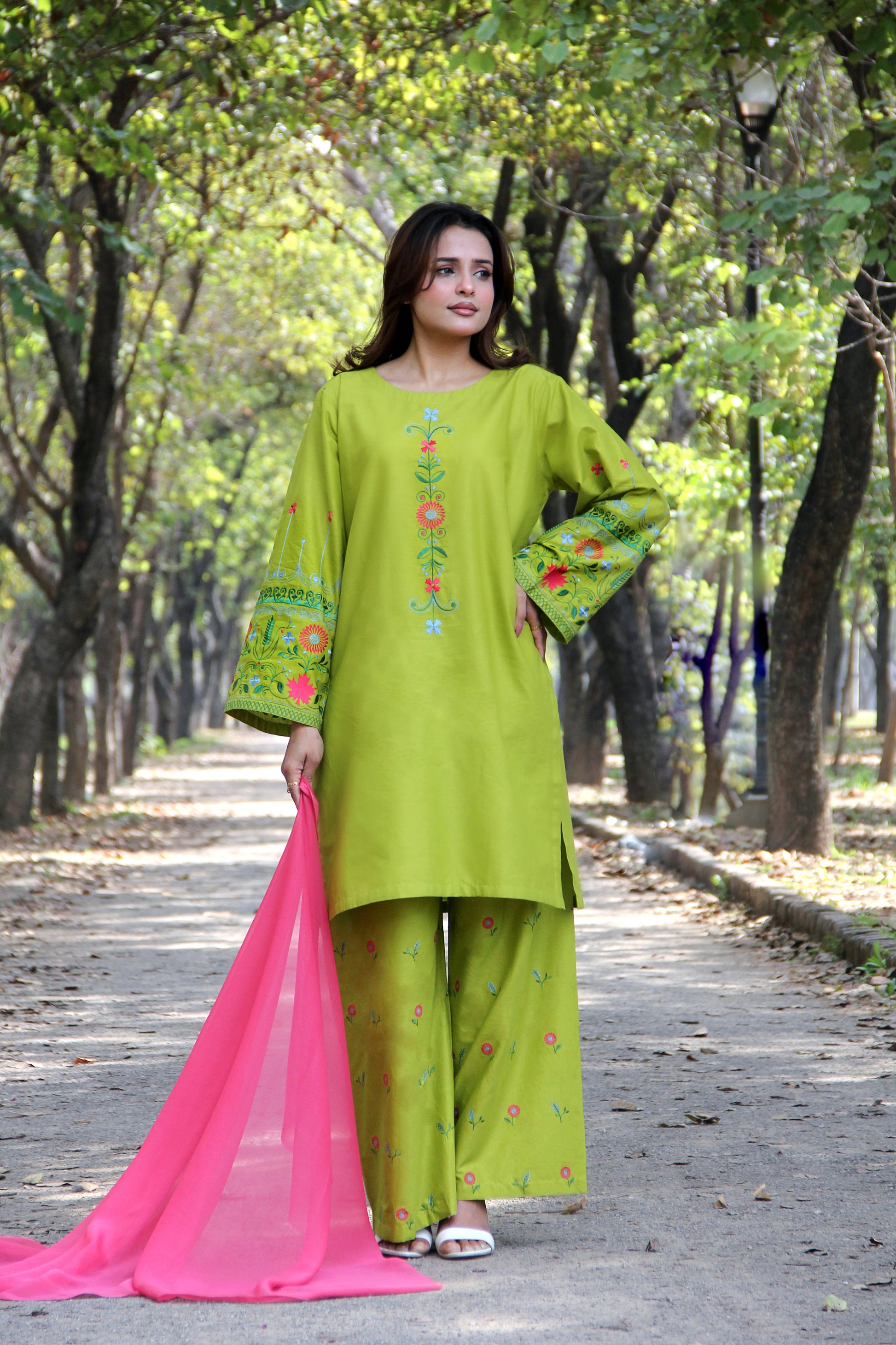 Woman in a green traditional outfit with pink dupatta standing in a park.