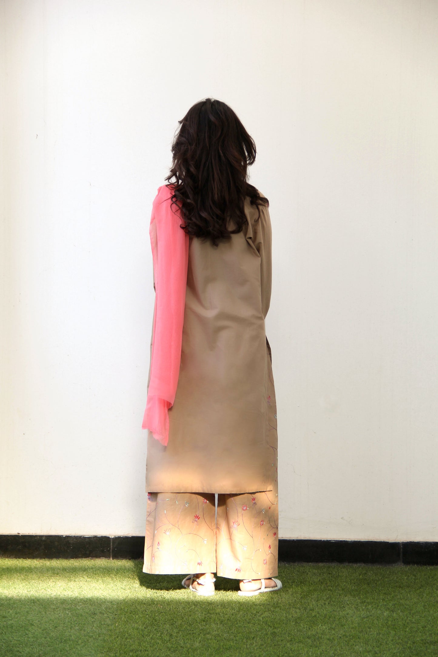 Person wearing a long beige coat with a pink sweater, standing on grass against a white wall.